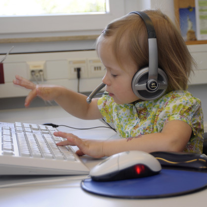 UK-Netzwerk Aufnahme eines blonden Kindes mit einem Logitech-Headset auf dem Kopf, welches an einer weißen Tastatur sitzt. Das Kind ist mitten in der Bewegung des Tippens und wirkt sehr konzentriert.